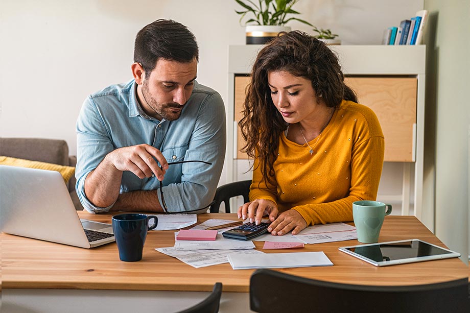 Foto de um casal verificando papeis e olhando a calculadora em uma mesa, fazendo contas