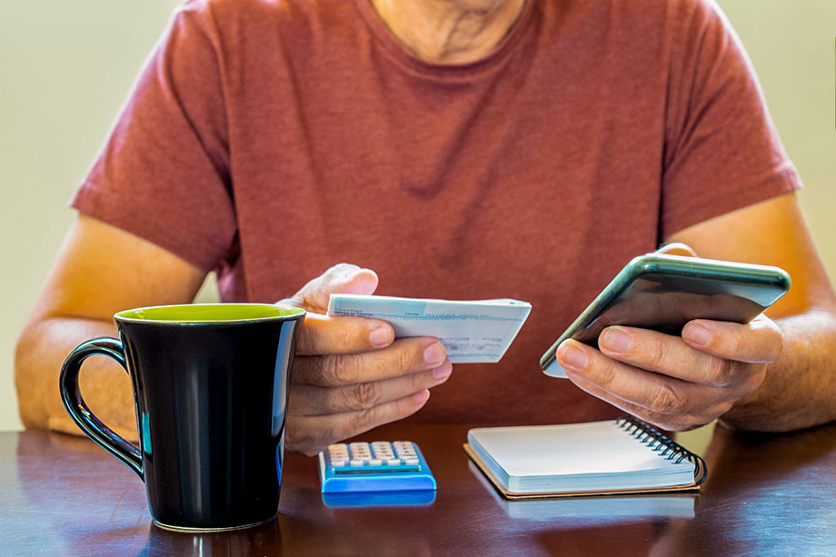 Foto mostra mão de homem segurando boletos e olhando celular, como se estivesse fazendo contas de dívidas